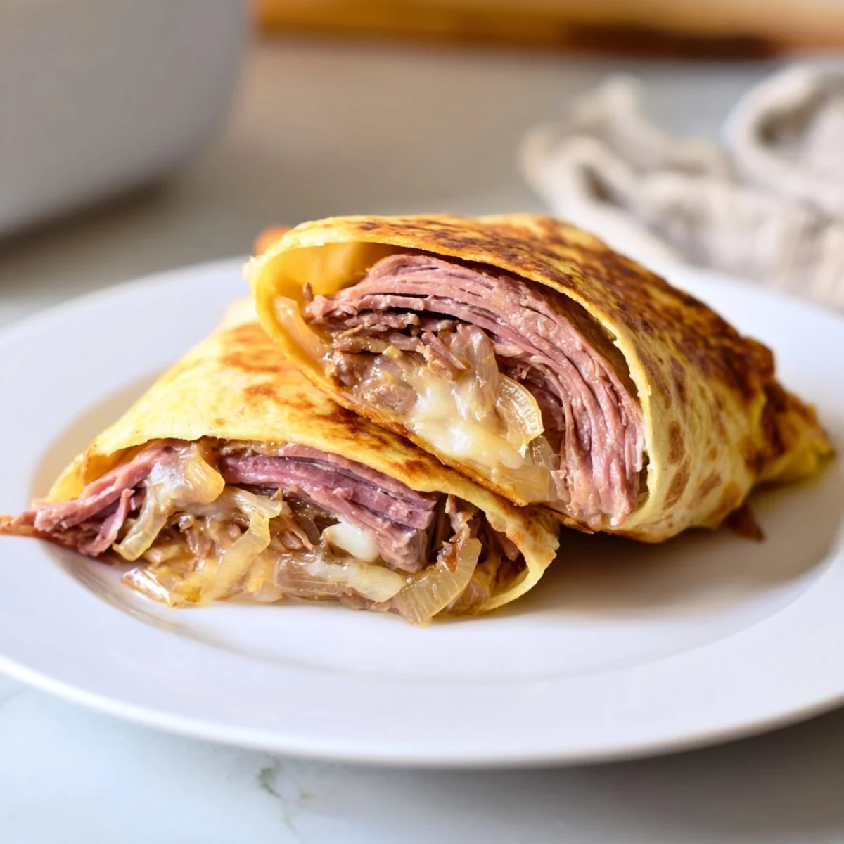 Golden brown Easy French Dip Tortilla Roll Ups sit on a plate, sliced in half to reveal layers of roast beef, melty provolone, and caramelized onions, with a small bowl of warm au jus nearby.