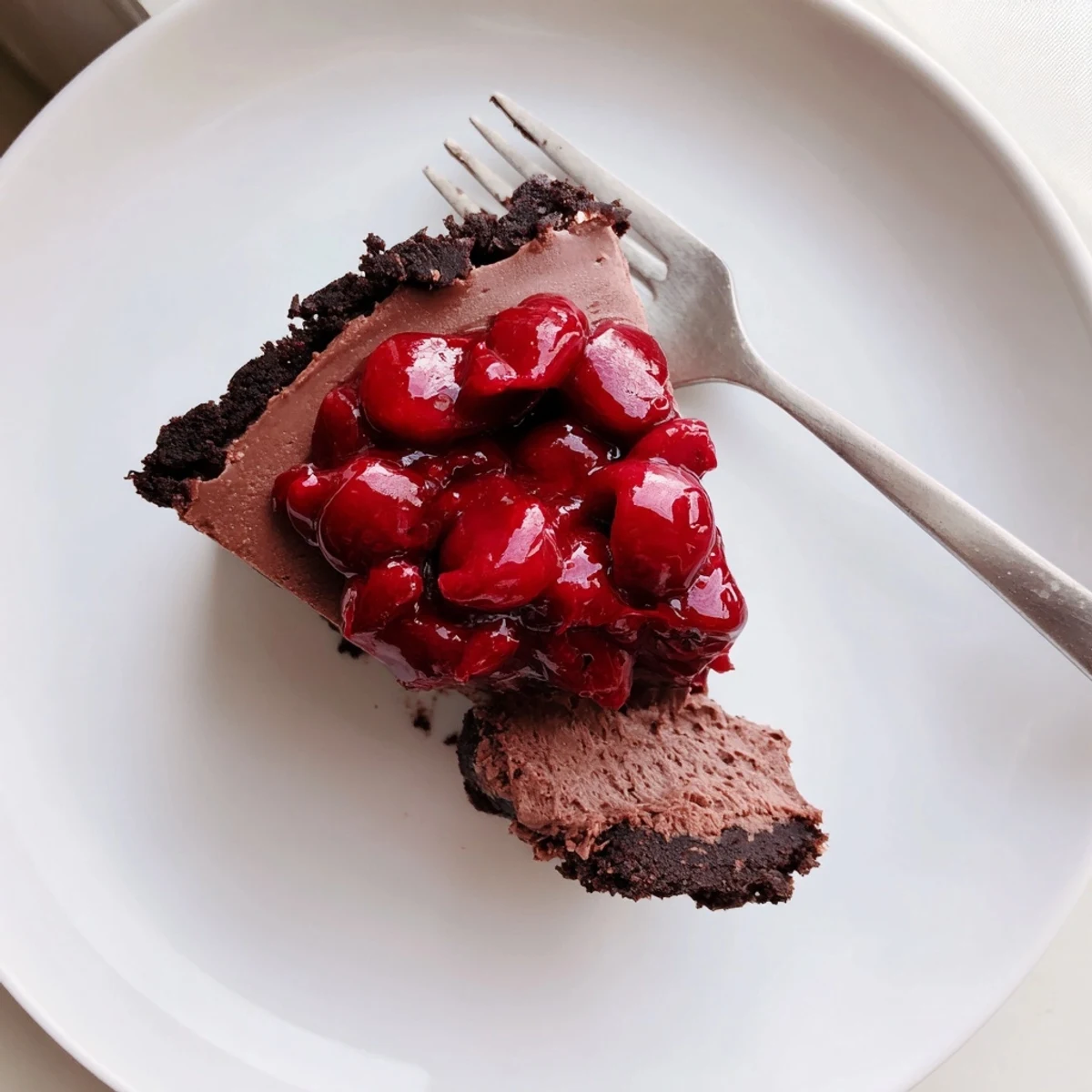 Rich dark chocolate cherry cheesecake slice topped with glossy red cherries on white plate