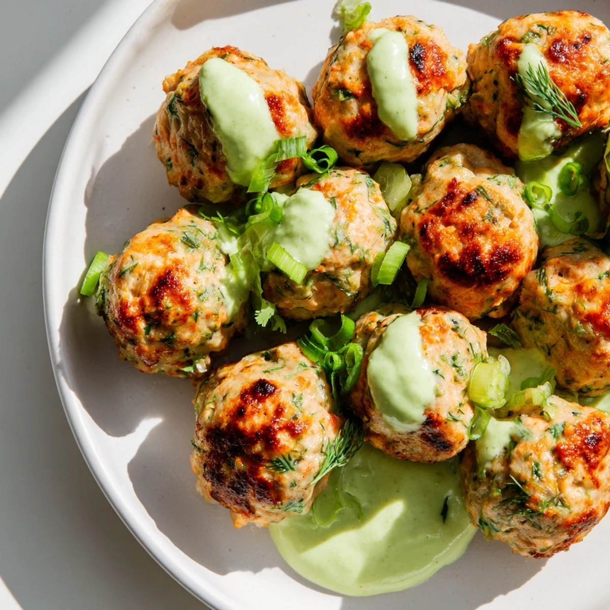 Golden baked salmon meatballs arranged on a white plate with vibrant green avocado sauce drizzled over the top