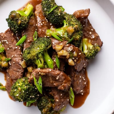 A close-up shows Better Than Takeout Beef With Broccoli served over fluffy white rice, garnished with green onions and sesame seeds.